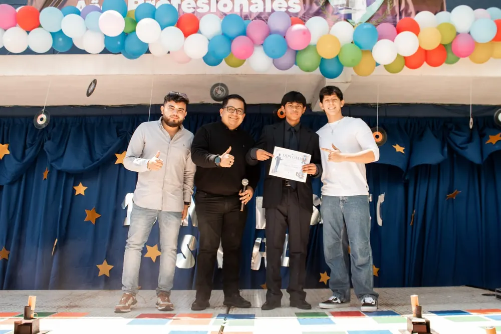 Estudiante recibiendo un diploma en la tarima junto a tres invitados, con globos de colores